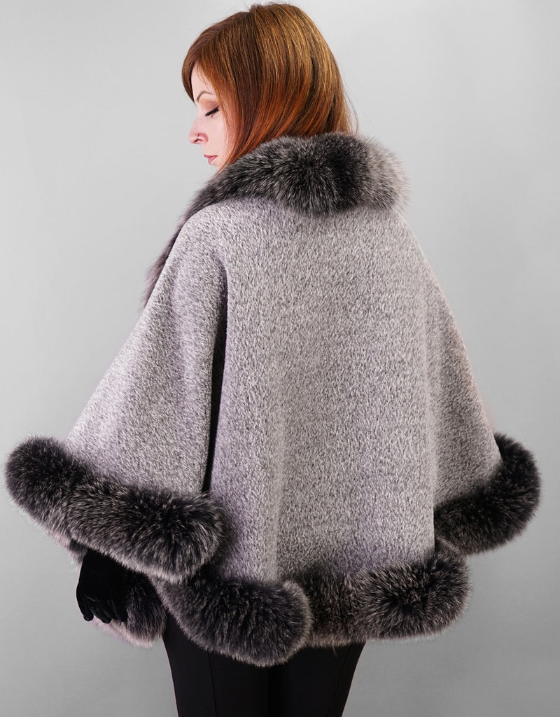Back view: A red haired woman is wearing a medium length silver cape with black with white tips Fox trim and black leather gloves