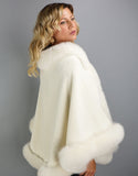 Side view: A blonde woman with green eyes is wearing a medium length off white cape. Medium length reaches mid thigh to knee. The cape has velvetty looking woven Alpaca wool with fluffy shiny fox trim